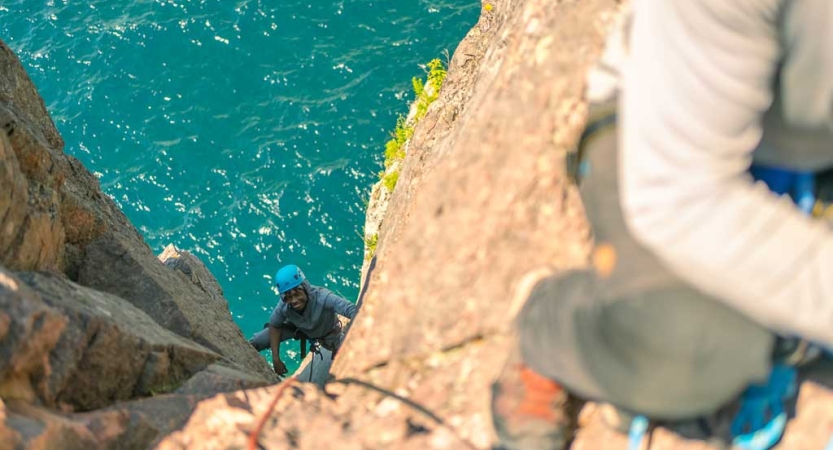 A person wearing safety gear is secured by ropes as they look up at the camera and smile while rock climbing. There is blue water below them. 